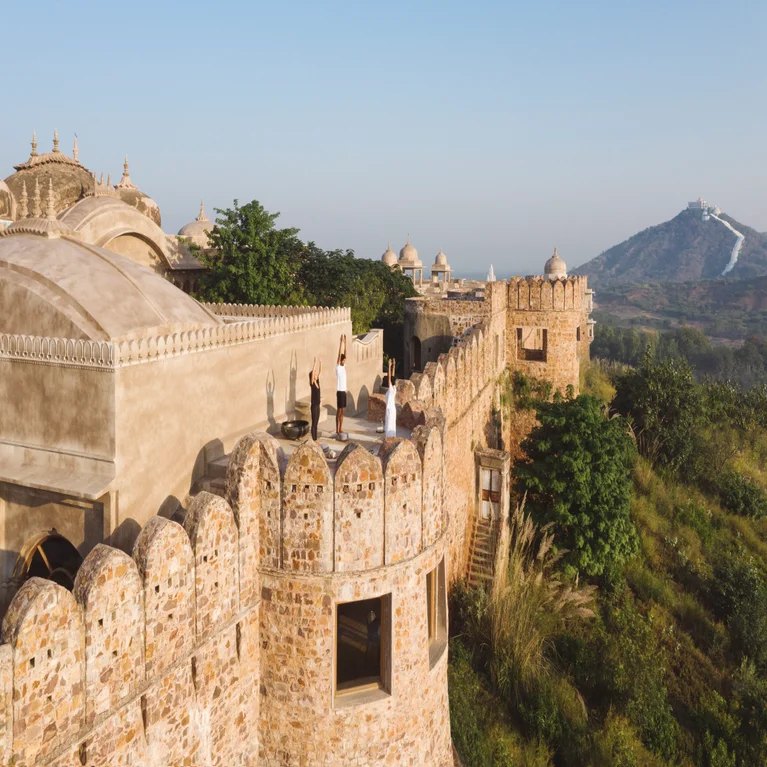 Stone fort with domed rooftops, stone walls and visitors, surrounded by lush greenery and distant hills beneath a blue sky.