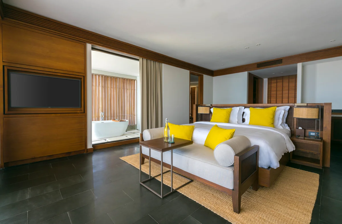A Sky Pool Suite bedroom, a king bed with yellow cushions on a woven rug, dark stone floor and a bathtub by the window