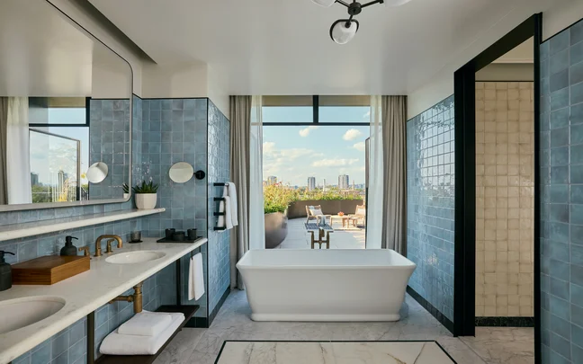 Salle de bains avec carrelage bleu, deux lavabos, baignoire à remous et douche à l'italienne. Une grande fenêtre offre une vue sur l'horizon de la ville et sur la terrasse extérieure sous des nuages épars.