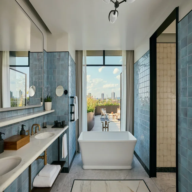 Bathroom with blue tile, twin sinks, soaking tub, and walk-in shower. Large window frames views of city skyline and outdoor terrace beneath scattered clouds.
