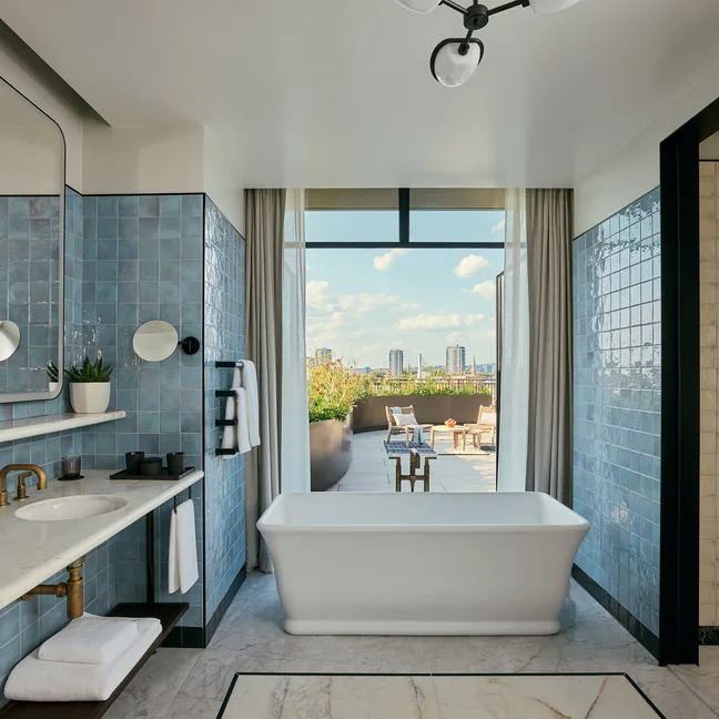 Bathroom with blue tile, twin sinks, soaking tub, and walk-in shower. Large window frames views of city skyline and outdoor terrace beneath scattered clouds.