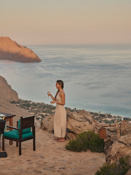 Los invitados cenan en una mesa situada en lo alto de un acantilado mientras el sol se pone sobre el océano y las montañas, rodeados de vistas costeras que realzan el entorno natural.