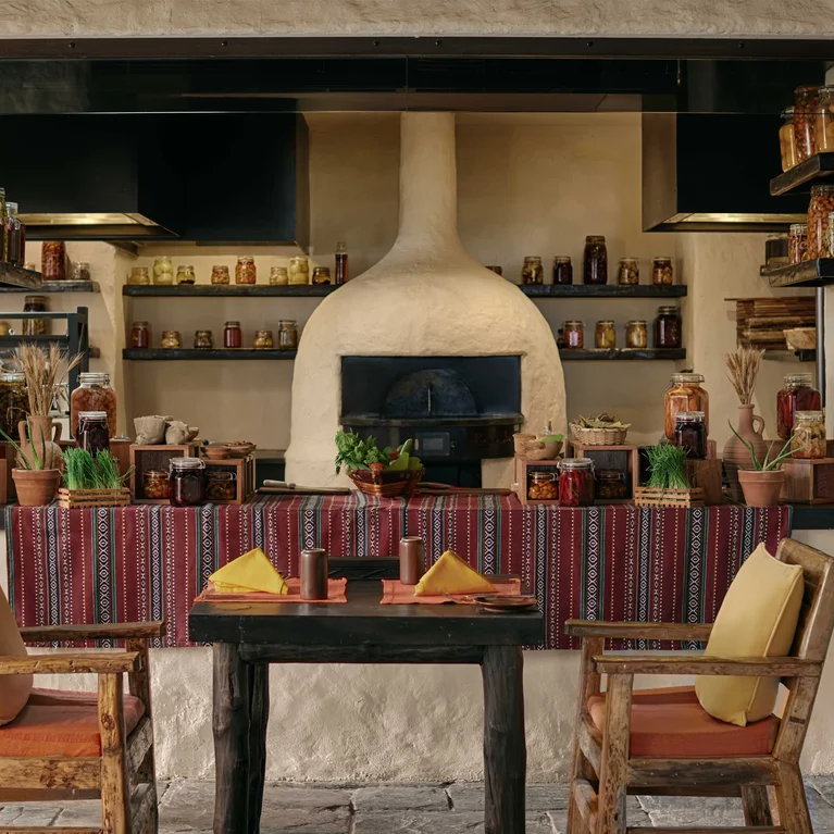 مطبخ مشرق يتميز بفرن قبة بيضاء، ورفوف مفتوحة مع برطمانات زجاجية، وطاولة مزينة بمناديل صفراء وكراسي خشبية مبطنة بمفارش مائدة منقوشة.