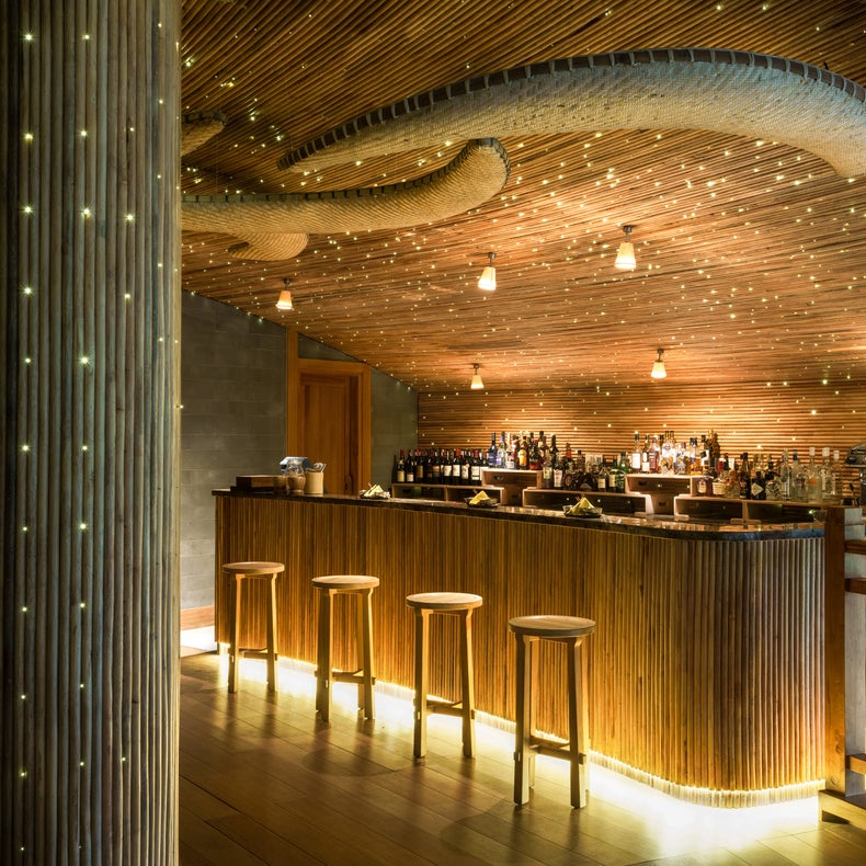 Bar area featuring stools and a gently curved wooden counter, with wavy wood ceiling panels and warm lighting highlighting natural materials.