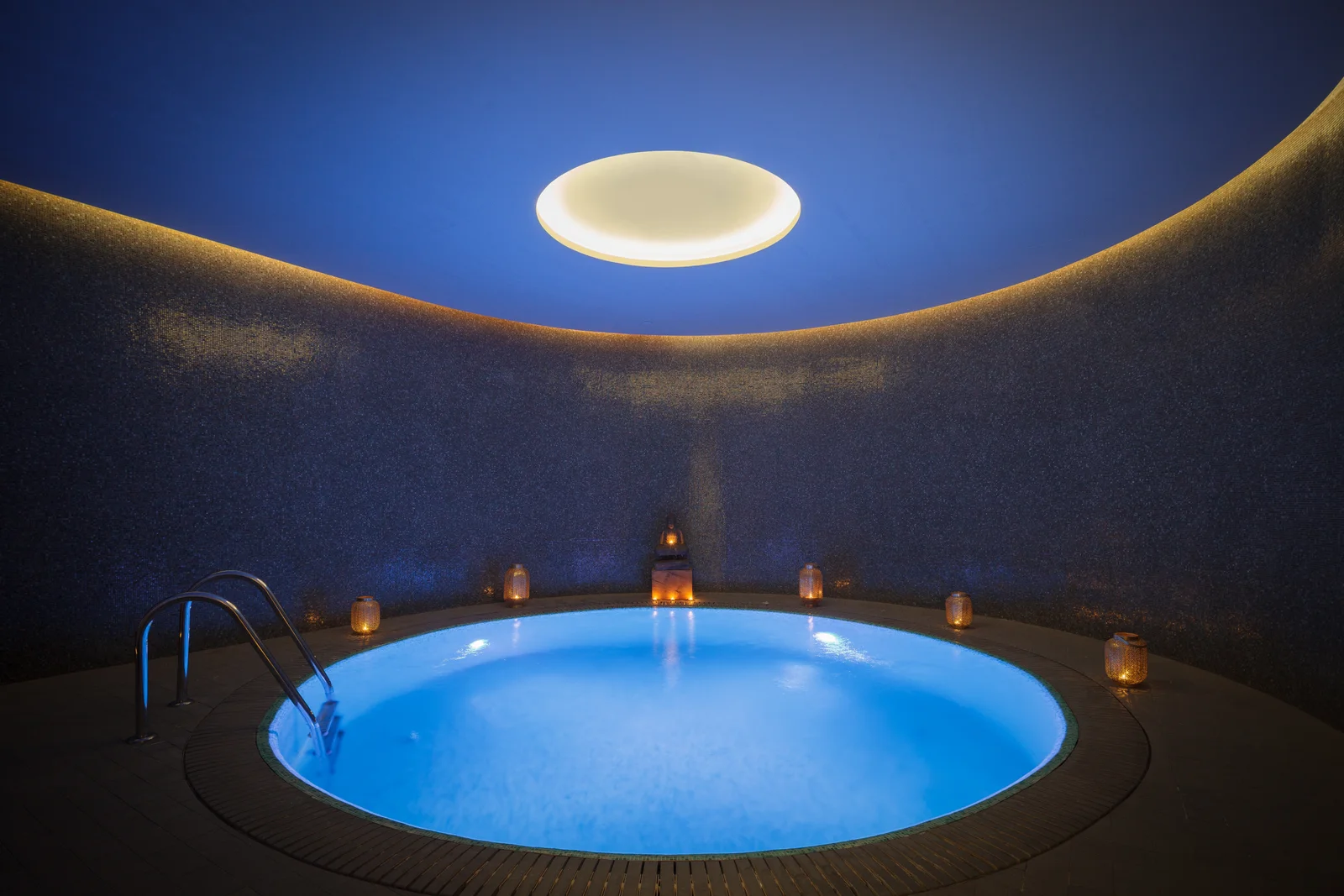 A round pool in a dark blue mosaic chamber, lit by a single circular light in the ceiling and candles set around the water's edge.