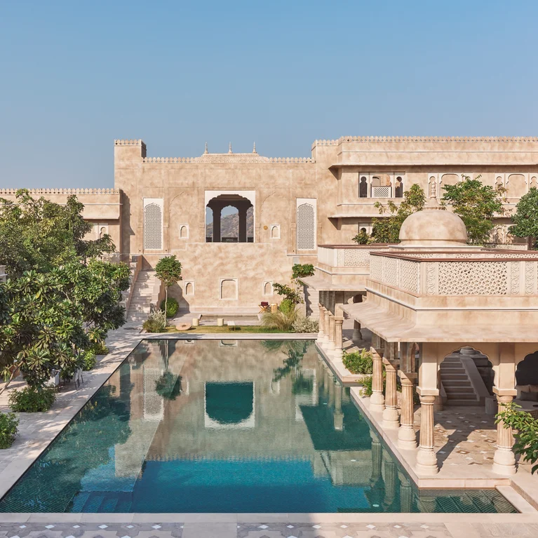 Beige stone architecture with detailed carvings encircles a large rectangular pool, surrounded by native plants beneath an open blue sky.