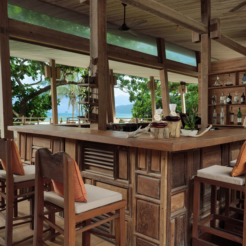 Wood bar with padded stools, shelves of beverages, and a backdrop of tropical greenery and ocean views, designed for comfort in a natural setting.