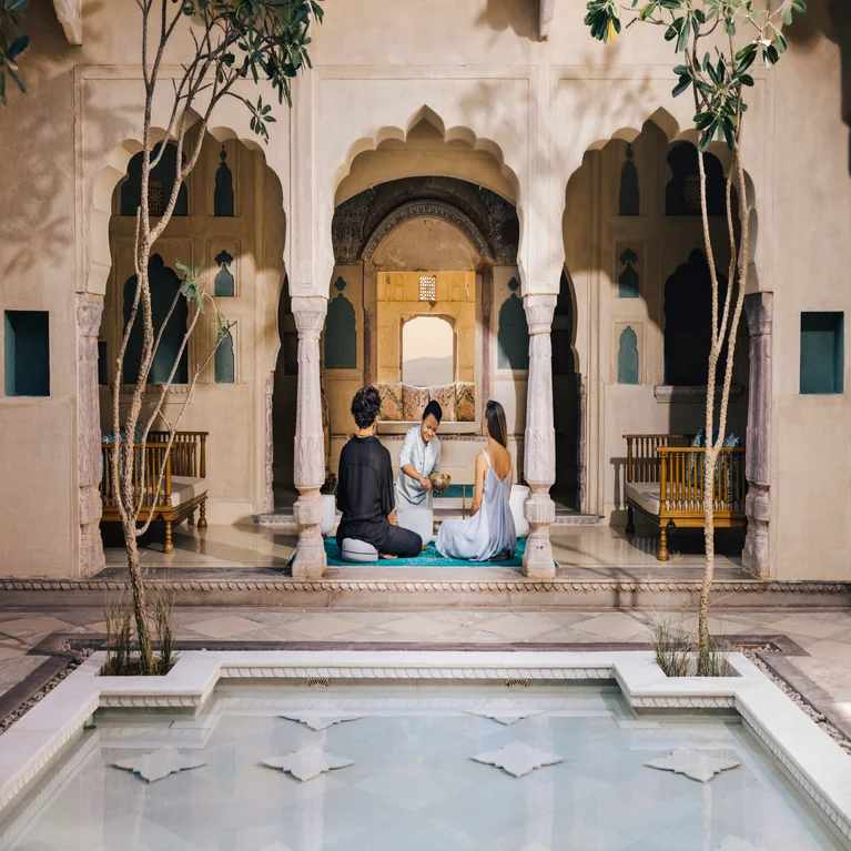 Guests enjoy a tranquil courtyard with arched columns, beside a decorative pool framed by detailed architecture and lush greenery.