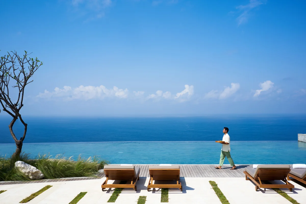 A host carrying a tray past the infinity pool, three loungers in front and nothing but sea and sky beyond