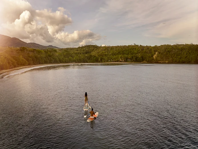 A guest enjoys paddleboarding in calm water near a green shoreline, under a sky with scattered clouds. White dress contrasts softly with the serene natural setting.