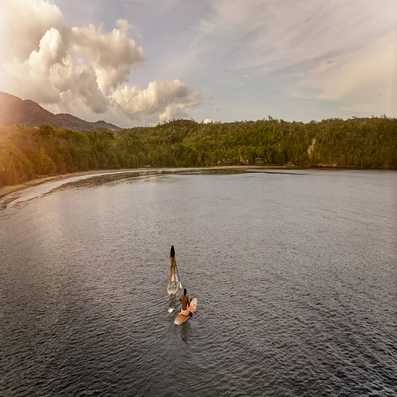 A guest enjoys paddleboarding in calm water near a green shoreline, under a sky with scattered clouds. White dress contrasts softly with the serene natural setting.
