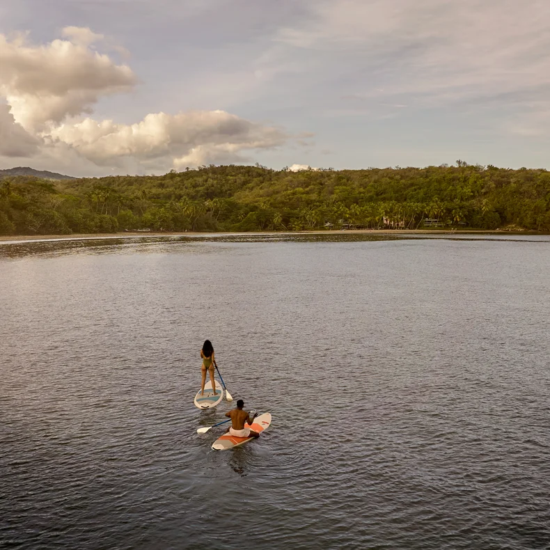A guest enjoys paddleboarding in calm water near a green shoreline, under a sky with scattered clouds. White dress contrasts softly with the serene natural setting.