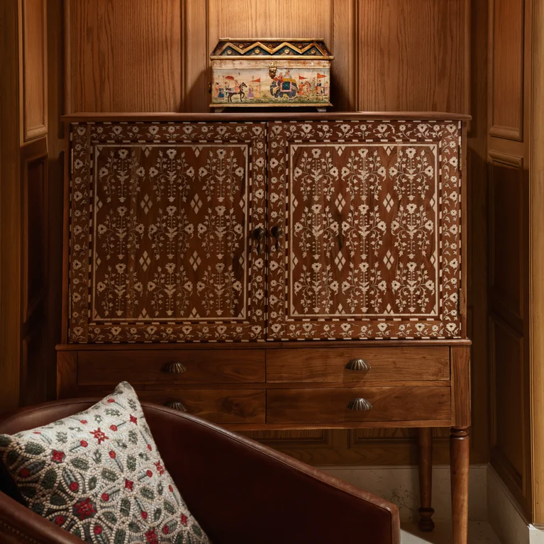 Cabinet with detailed white inlay sits by a wood-paneled wall, topped with a patterned box; a brown leather chair and cushion placed thoughtfully in front.