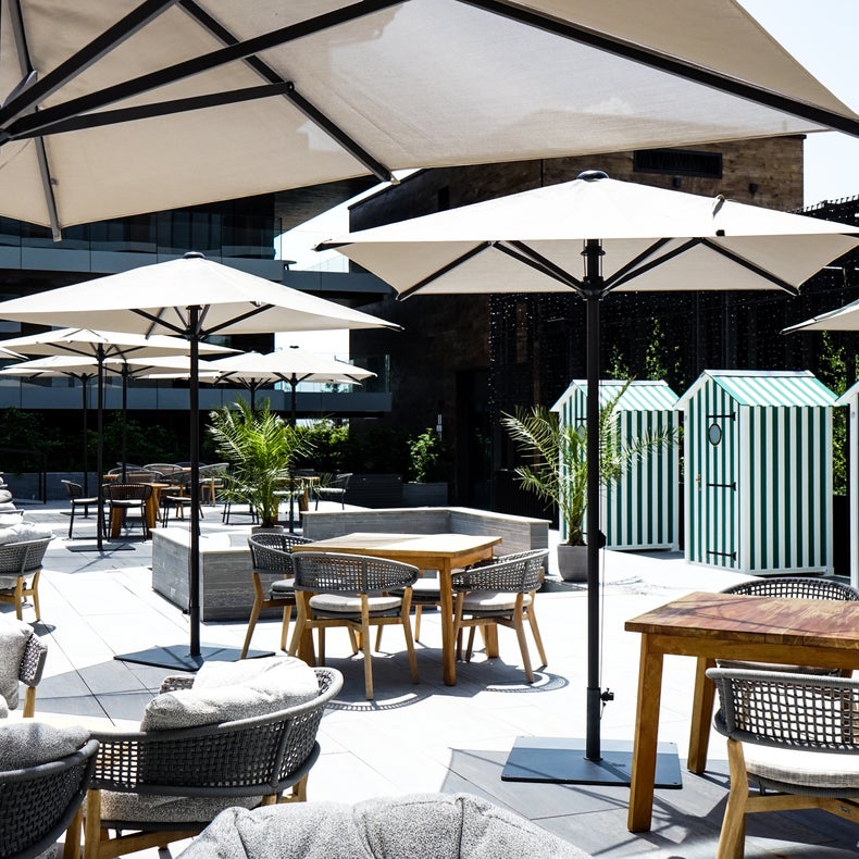 Tables and chairs shaded by large umbrellas, with striped cabanas behind. Potted plants introduce greenery to a sunlit, contemporary space.