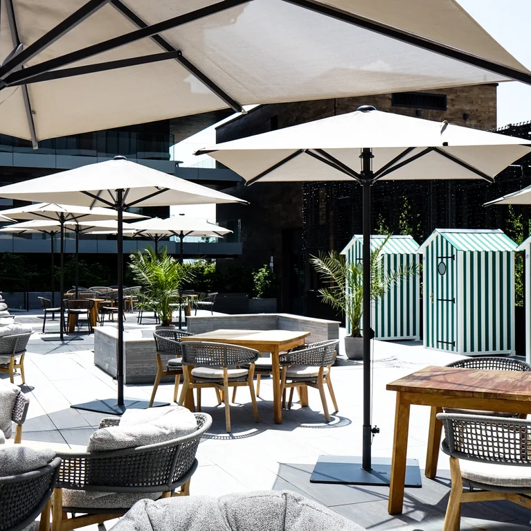 大型遮阳伞遮住了桌椅,后面是条纹小屋。盆栽植物为这个充满阳光的现代空间增添了绿意。.