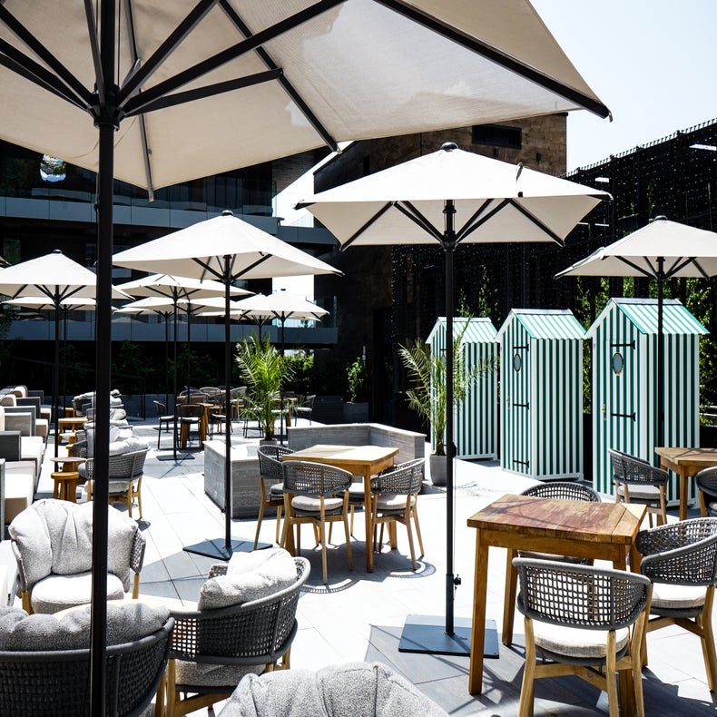 Tables and chairs shaded by large umbrellas, with striped cabanas behind. Potted plants introduce greenery to a sunlit, contemporary space.