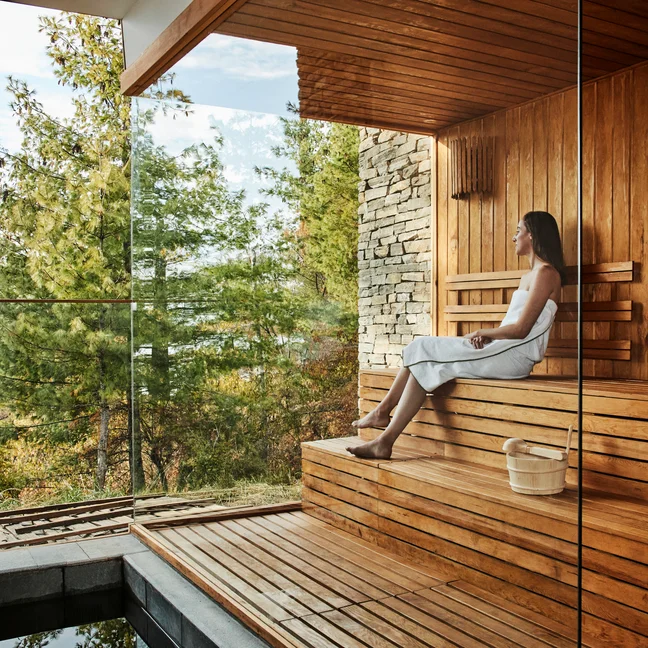 El huésped disfruta de una sesión de sauna en unos cómodos bancos de madera, con amplios ventanales que ofrecen unas tranquilas vistas de los árboles y el paisaje natural que lo rodean.
