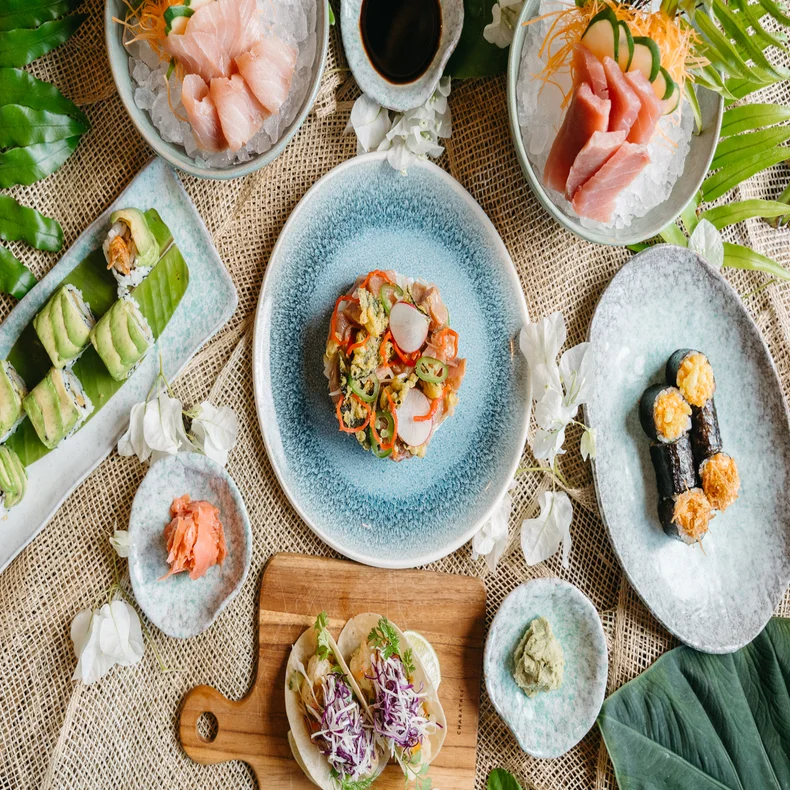 Sushi, sashimi, and rolls are presented on plates with green leaves and edible flowers; dishes sit on a textured table for an inviting dining experience.
