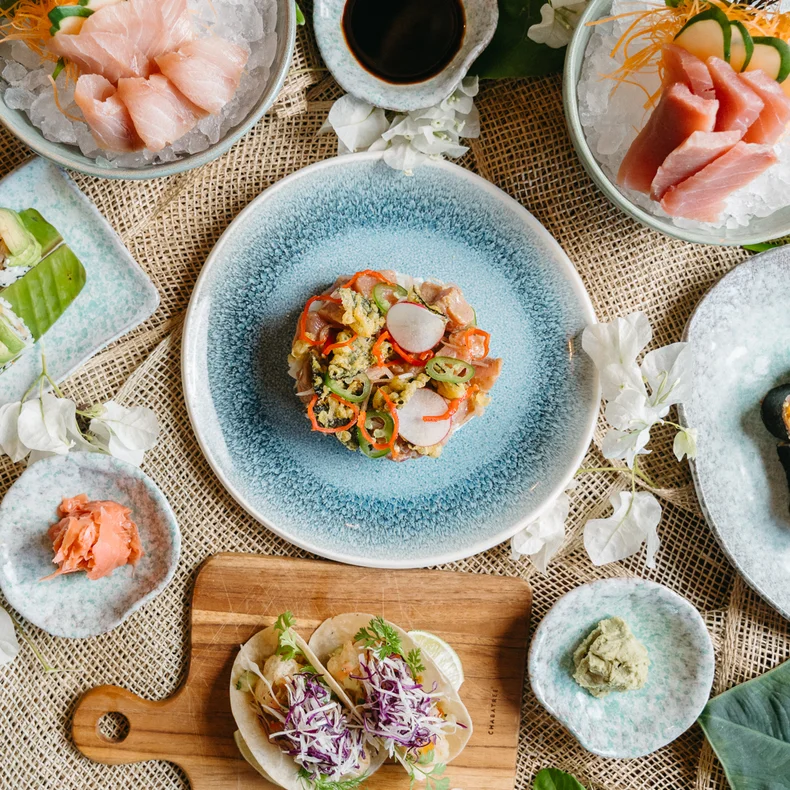 Sushi, sashimi, and rolls are presented on plates with green leaves and edible flowers; dishes sit on a textured table for an inviting dining experience.