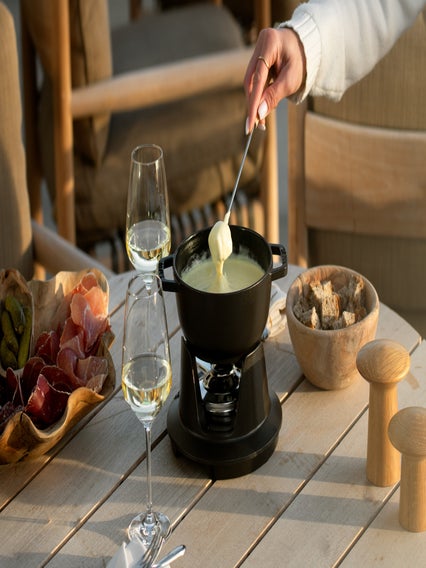 Bread is dipped into cheese fondue at a wooden table arranged with wine, charcuterie, and extra bread in a warm, inviting dining space.