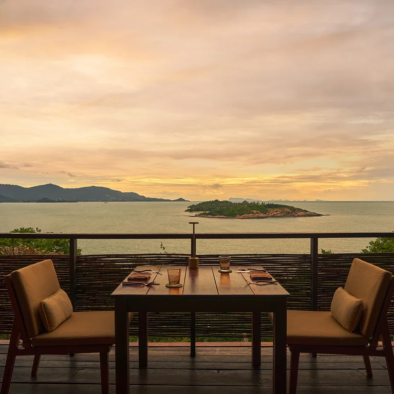 Mesa de comedor para dos dispuesta en un balcón con serenas vistas al mar, una pequeña isla a lo lejos y montañas bajo el cielo del atardecer.