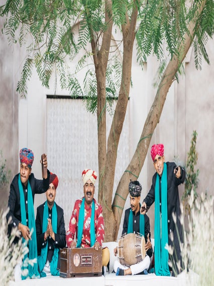 Musicians in traditional Rajasthani clothing perform with a harmonium and dholak drum in a garden setting.