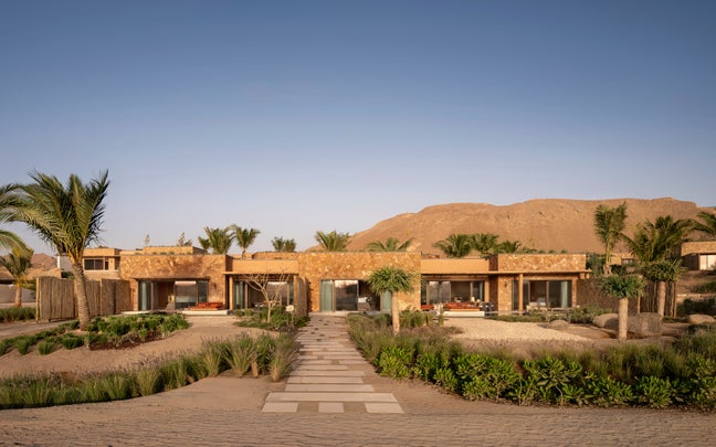 Single-story villa with expansive windows, shaded by palm trees and greenery, connected by a stone path. Set in a desert landscape with a mountain rising behind.