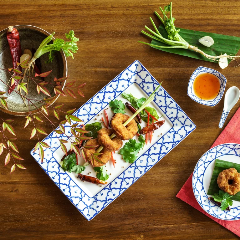 Fried dough rings on banana leaves, topped with fresh herbs, served next to dipping sauce and salad on a wooden table set with a red napkin.