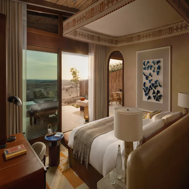 Spacious hotel room featuring a king bed, natural materials, work desk, and sliding doors to a patio with lounge chairs overlooking the desert at sunset.