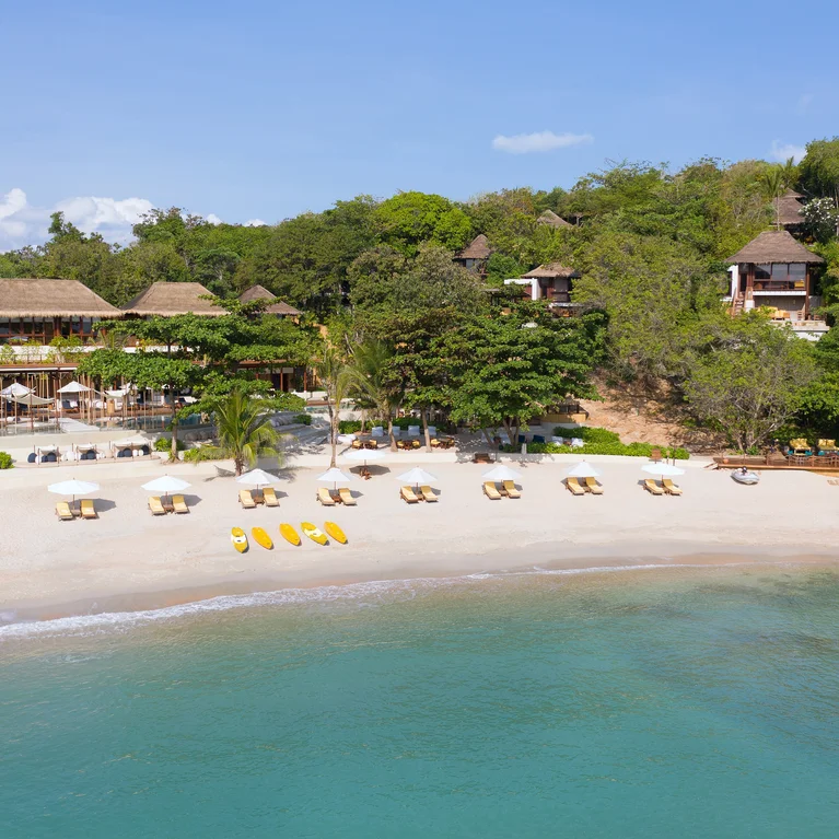 فيلات ذات أسقف من القش تقع على طول الرمال البيضاء مع كراسي استلقاء للتشمس ومظلات وزوارق الكاياك. تلتقي المياه الفيروزية الصافية مع النباتات المورقة على الشاطئ.