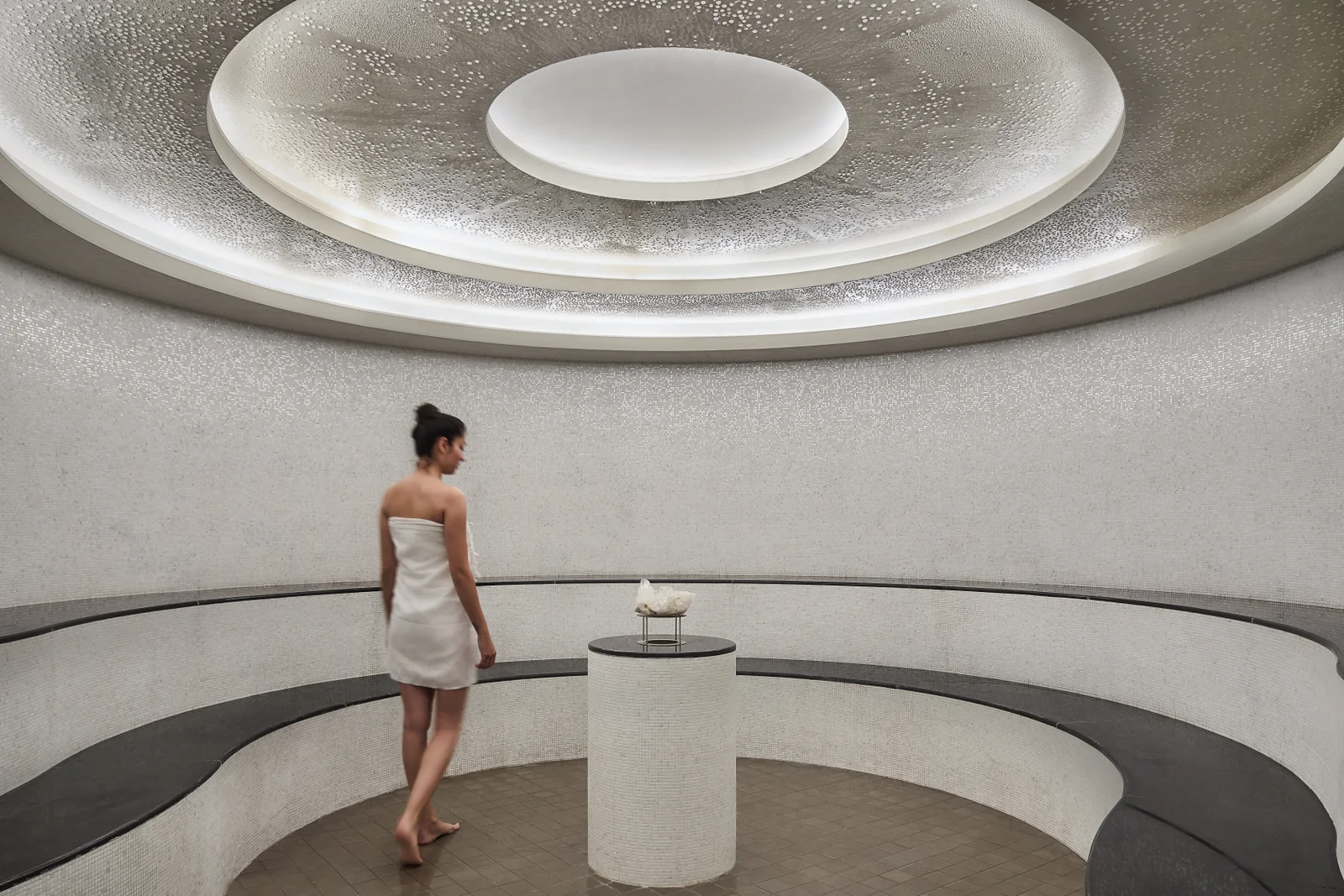 A domed white room with a circular skylight ring above and a curved bench running around the wall, a single guest crossing the floor.