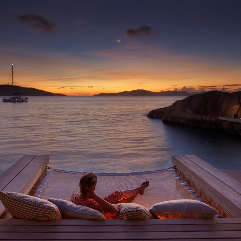 Los invitados se sientan en asientos de red sobre el agua al atardecer, rodeados de almohadas y farolillos; en el horizonte se ven un barco y unas colinas lejanas.