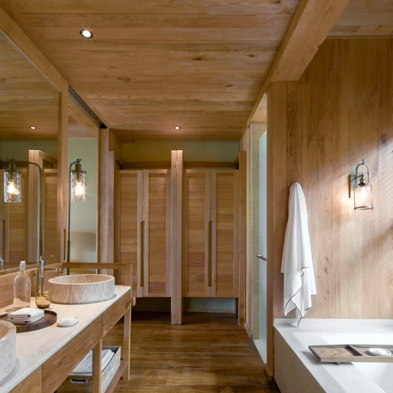 Bathroom features wood paneling, stone sinks, spacious mirror, a towel by the bathtub, and essentials arranged along the tub for a calming space.