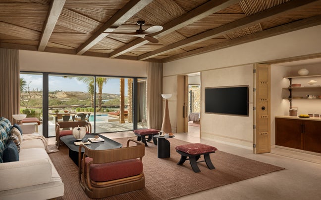 Serene lounge featuring natural wood ceiling, low-profile seating, open shelves, and glass doors opening to a pool bordered by palm trees.