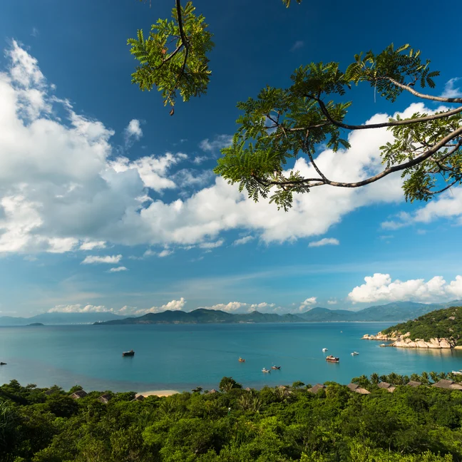 Clear blue water with boats floats beside green hills and tree branches; gentle clouds drift across the sky, reflecting a tranquil coastal setting.