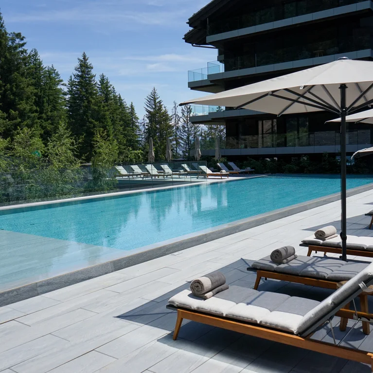 宁静的泳池边摆放着躺椅和遮阳伞,绿树成荫,眼前是一座现代建筑,头顶是蔚蓝的天空。.