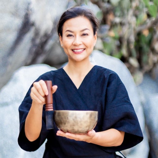 A woman in a black robe holds a brass singing bowl and striker outdoors, surrounded by natural rocks and greenery, connecting with the environment.