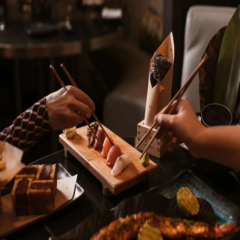 Diners use chopsticks to lift nigiri sushi from a wooden board.