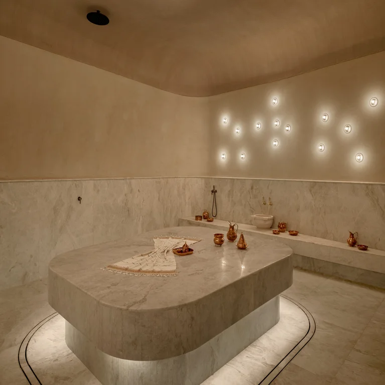 Marble Turkish bath with gentle lighting, copper bowls, and towels arranged on a central stone slab. A tranquil space designed for relaxation and wellbeing.