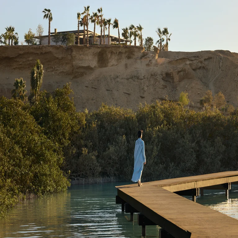 Guest in a robe on a wooden boardwalk over water, surrounded by lush greenery and a rocky hillside with palm trees in the distance.