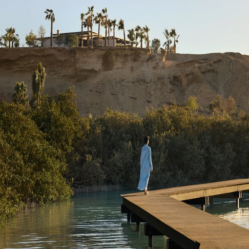Guest in a robe on a wooden boardwalk over water, surrounded by lush greenery and a rocky hillside with palm trees in the distance.