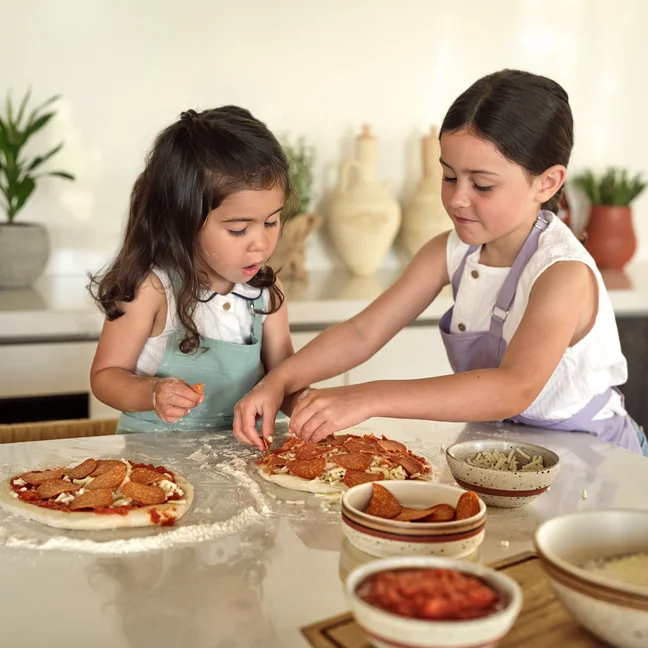 Children in aprons prepare pizza, arranging toppings on dough at a sunlit kitchen counter with fresh ingredients, reflecting a mindful culinary experience.