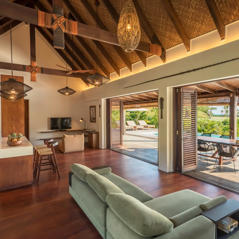 别墅起居区设有高高的木质天花板、绿色沙发、开放式厨房以及通往池畔露台的宽大落地门,可欣赏花园和水景。.