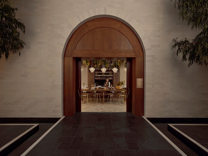 An arched entrance opens to a warmly lit dining area with wooden tables, greenery overhead, and a staff member behind the counter.