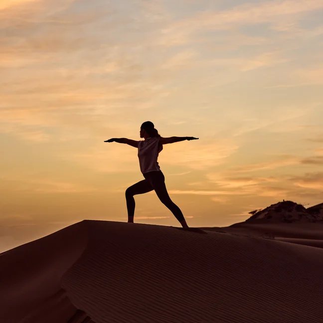 Someone in a yoga pose atop a sand dune as the sun sets, outlined against a sky painted with soft colors and scattered clouds.