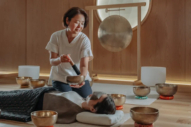 A wellness practitioner guides a singing bowl sound session in a tranquil wooden room, with a guest lying peacefully and eyes closed.