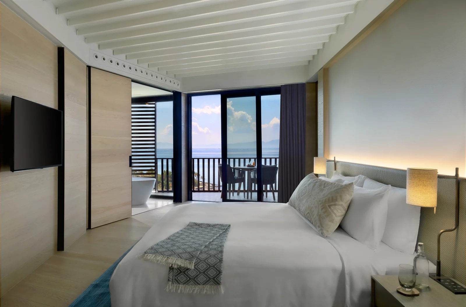 A sea-view room with pale timber walls and a slatted ceiling, the bed facing sliding doors that open onto a balcony above the water.