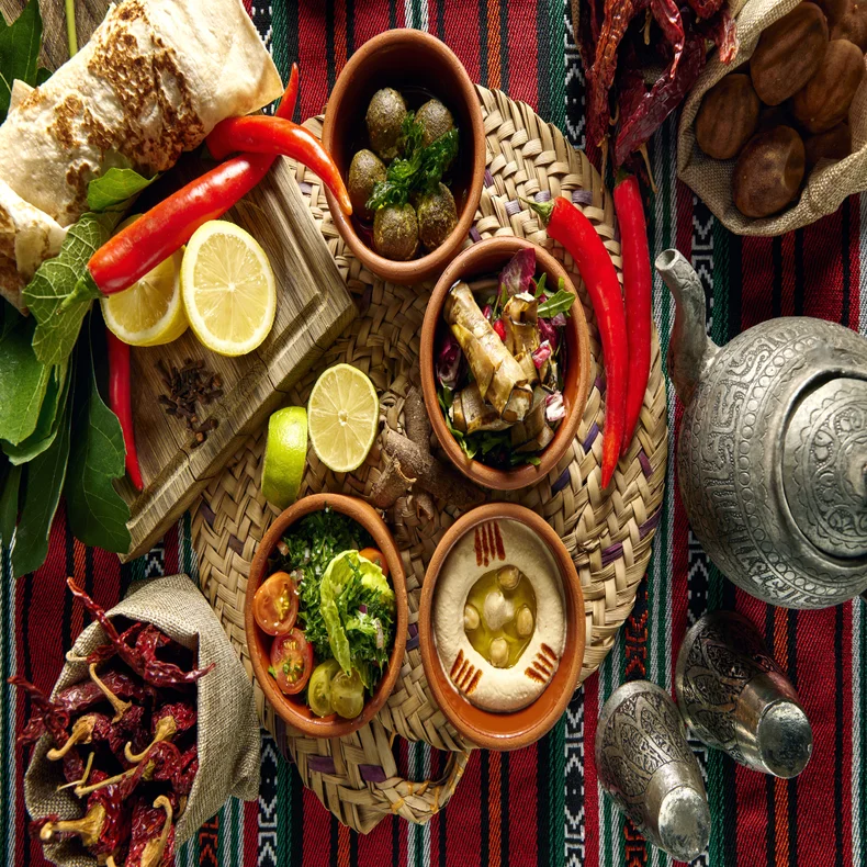 Middle Eastern cuisine, fresh vegetables, and bread set on a woven mat with a metal teapot atop a red patterned cloth. Warm, inviting presentation.