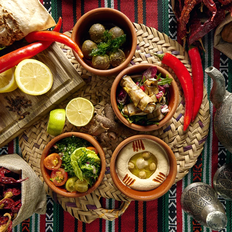 Middle Eastern cuisine, fresh vegetables, and bread set on a woven mat with a metal teapot atop a red patterned cloth. Warm, inviting presentation.