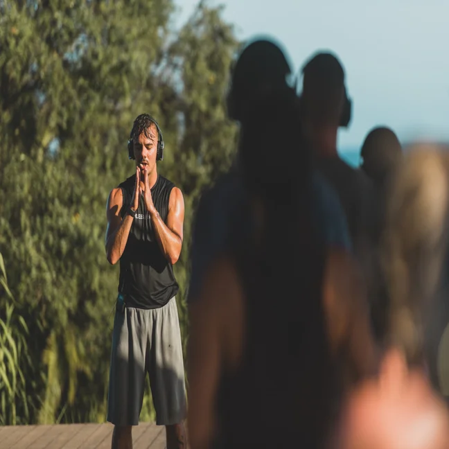 An instructor leads a mindful outdoor fitness session, hands together in front of his face. Participants join in, slightly out of focus in the foreground.